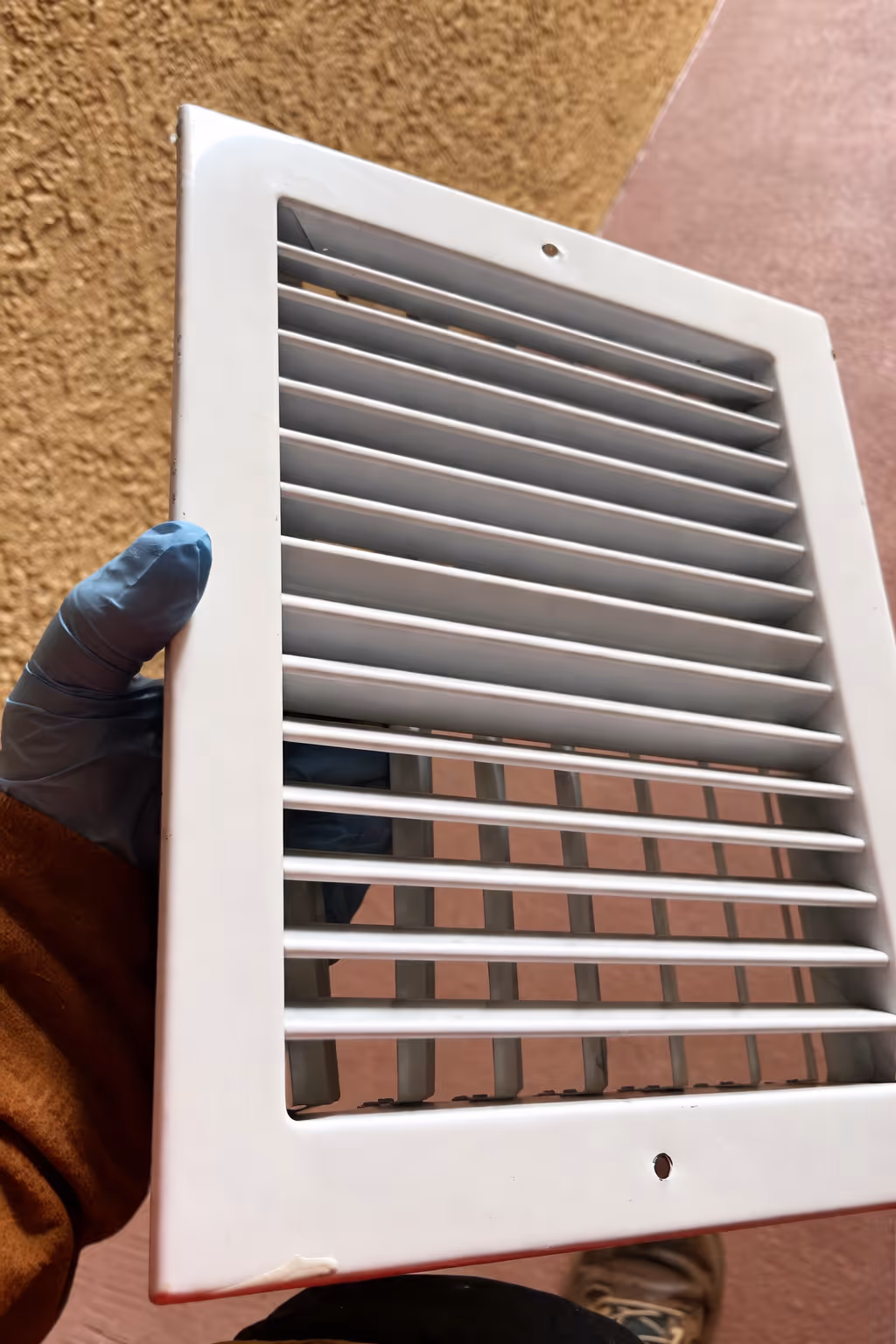 Hand in blue glove holding a white rectangular air vent cover with horizontal slats.